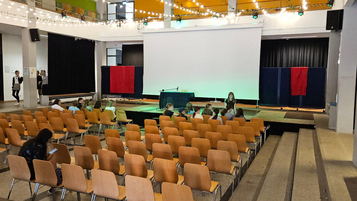 Die Aula einer Schule mit einer Bühne, auf der ein Tisch mit Mikro steht. Davor mehrere Stuhlreihen.